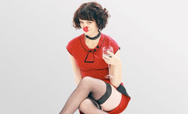 A woman in a red clown nose with short brown hair wearing a short red dress and black stockings holds a wine glass.