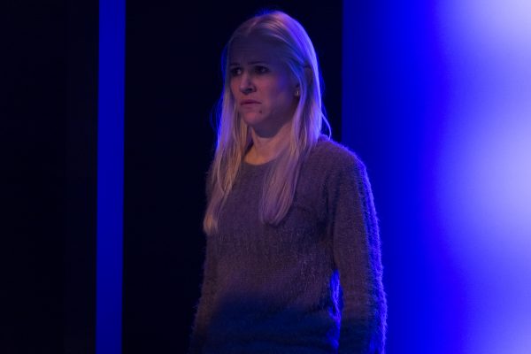 A woman stands in front of a glowing blue wall in a play