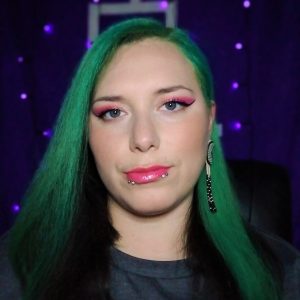 A close up of a woman with long, straight dyed green hair wearing bright pink lipstick and eyeshadow and piercings in her lip.