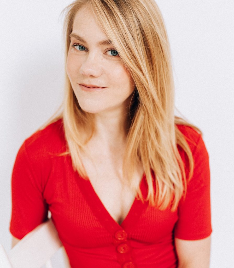 Amelia has blond hair and wears a red shirt. She is seated on a white wooden chair and looks out at you with a small grin, perhaps a bit impishly.