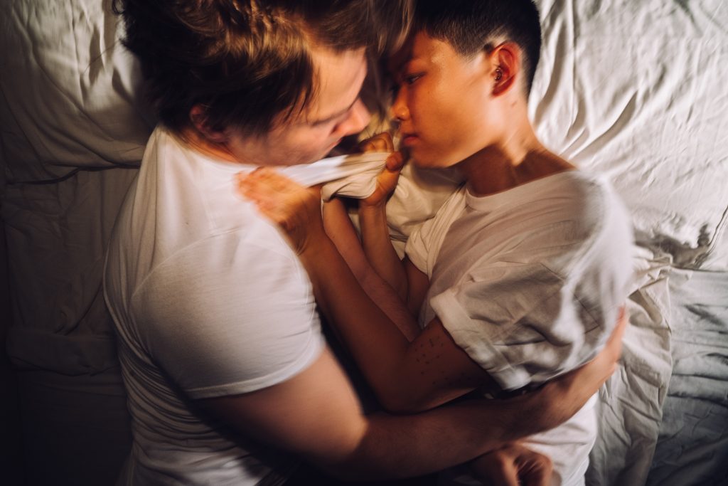 A man and a woman are in a bed of white sheets and they wear white t-shirts. The woman holds the man by the collar of his shirt aggressively.