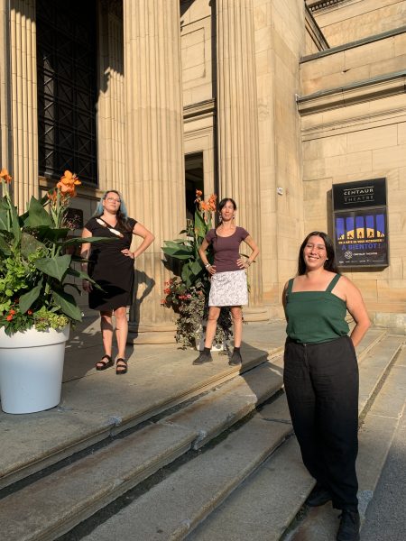 Three women stand on the stone steps in from of Centaur Theatre in summer